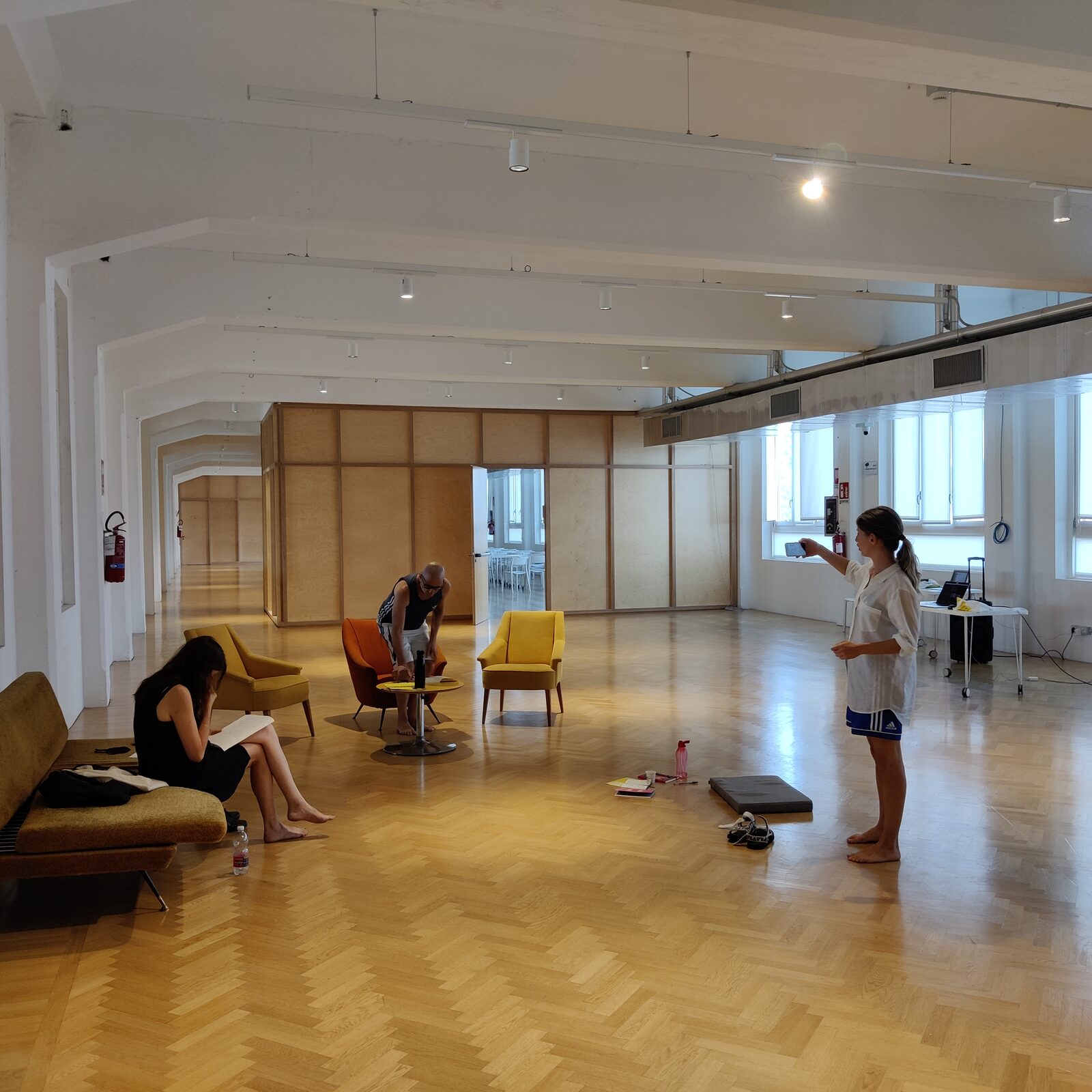 A near-empty dance studio, herringbone floor, three figures sitting still along the walls.